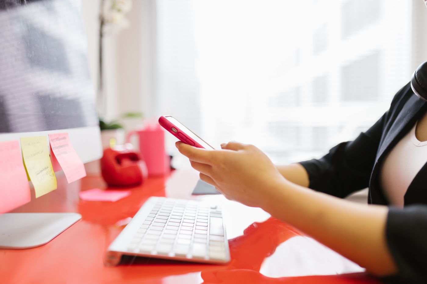 A person at a bright office desk holding a phone beside a keyboard and monitor